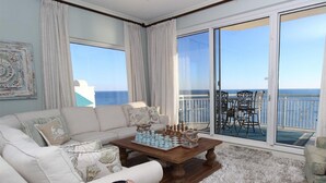 A living room with a large window overlooking the ocean, a chessboard on a coffee table, and a view of a deck with outdoor furniture. - Indigo East 2001 (Pensacola)