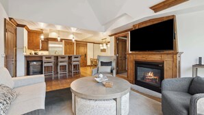 A spacious living area with a fireplace, a television mounted on the wall, and a kitchen in the background.