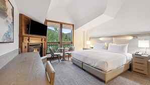 A hotel room with a large bed, a TV mounted on a wooden panel, a fireplace, a dining table with chairs, and a view of the outdoors through large windows.