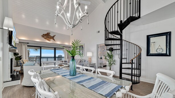 A spacious dining area with a glass table, white chairs, and a blue-striped runner. A chandelier hangs from the ceiling, and a spiral staircase is visible in the background. - Shipwatch C301 (Pensacola)