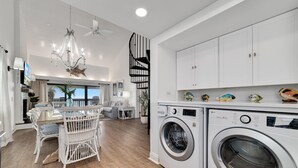 A modern kitchen with white cabinetry, a built-in washer and dryer, a chandelier, and a dining area with a white table and chairs. - Shipwatch C301 (Pensacola)