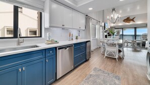 A modern kitchen with white and blue cabinets, a dishwasher, and a dining area with a view. - Shipwatch C301 (Pensacola)