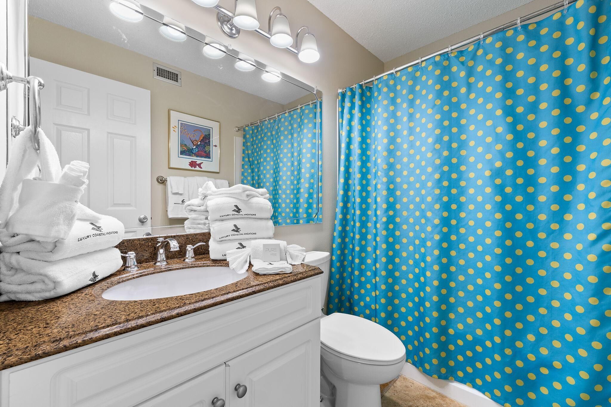 Een badkamer met een wastafel, spiegel en toilet. Er liggen handdoeken op het aanrecht en er is een blauw stippen douchegordijn.