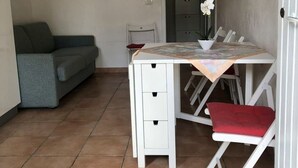 Una habitación pequeña y bien iluminada con una mesa y sillas blancas, un sofá gris, un cuadro en la pared y un televisor montado en la pared.