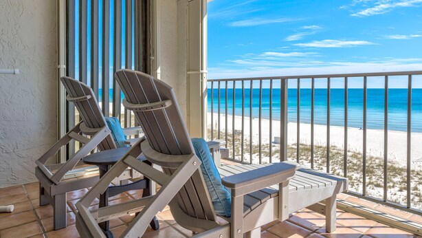 Un balcone con due poltrone e un tavolino, affacciato su una spiaggia con acqua cristallina e una battigia sabbiosa.