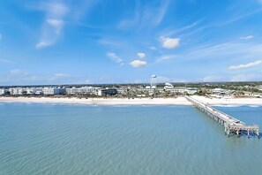 A coastal hotel with a pier extending into the water, beachfront rooms, and a clear sky.