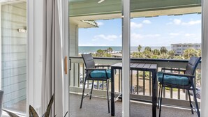 A balcony with two chairs and a table, overlooking a beach with palm trees and a clear sky. - 1 BR Top Floor Isle of Palms Oceanfront Condo | Pool | Free Wifi & Parking (Isle of Palms)