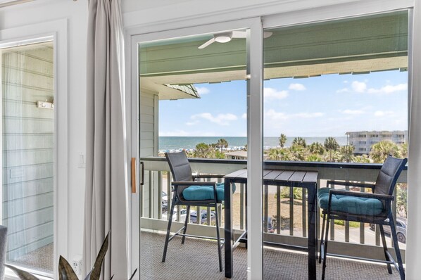 A balcony with two chairs and a table, overlooking a beach with palm trees and a clear sky.