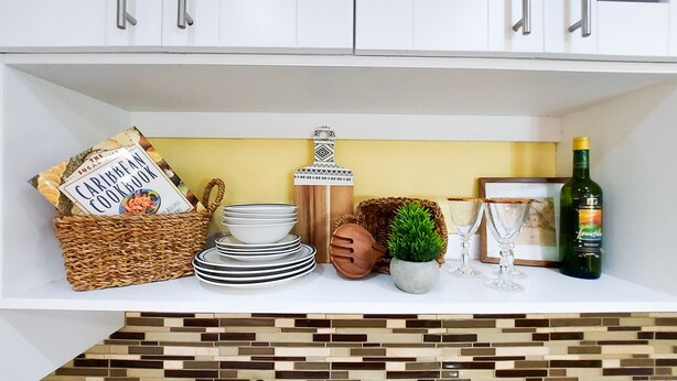 A kitchen shelf with a Caribbean cookbook, stacked plates, a decorative bottle, a small plant, and a bottle of wine.