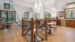 A spacious living area with a wooden staircase, large windows, and a kitchen with wooden cabinets.