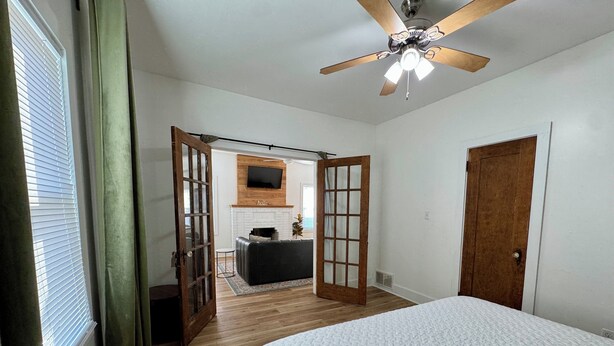 Une chambre à coucher avec un lit, un plancher en bois, un ventilateur de plafond, un téléviseur fixé au-dessus d’une cheminée et une porte donnant sur une autre pièce.
