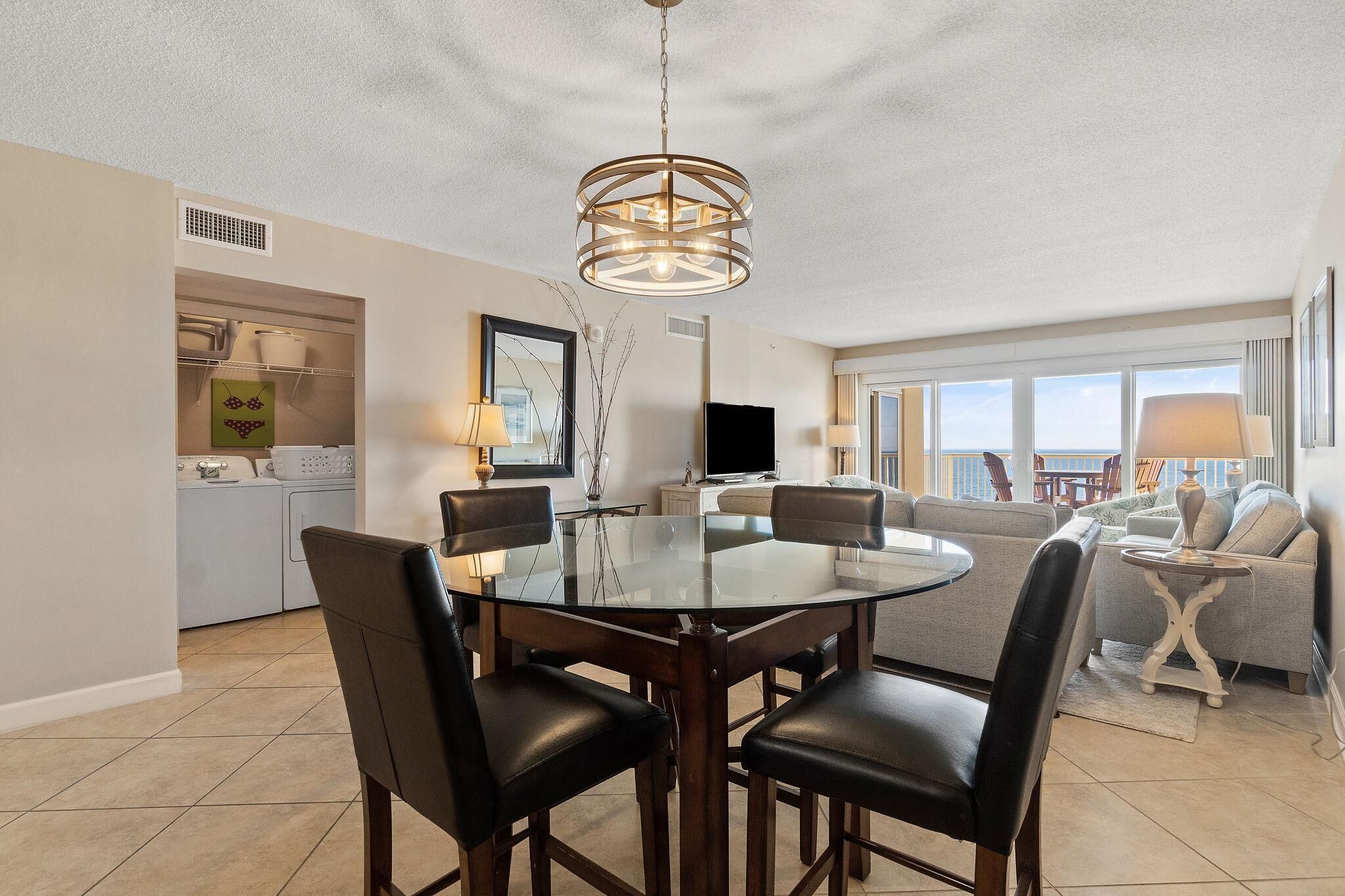 Un espace repas moderne avec une table ronde en verre et des chaises noires, un luminaire au plafond et une vue sur un balcon agrémenté d’un canapé.
