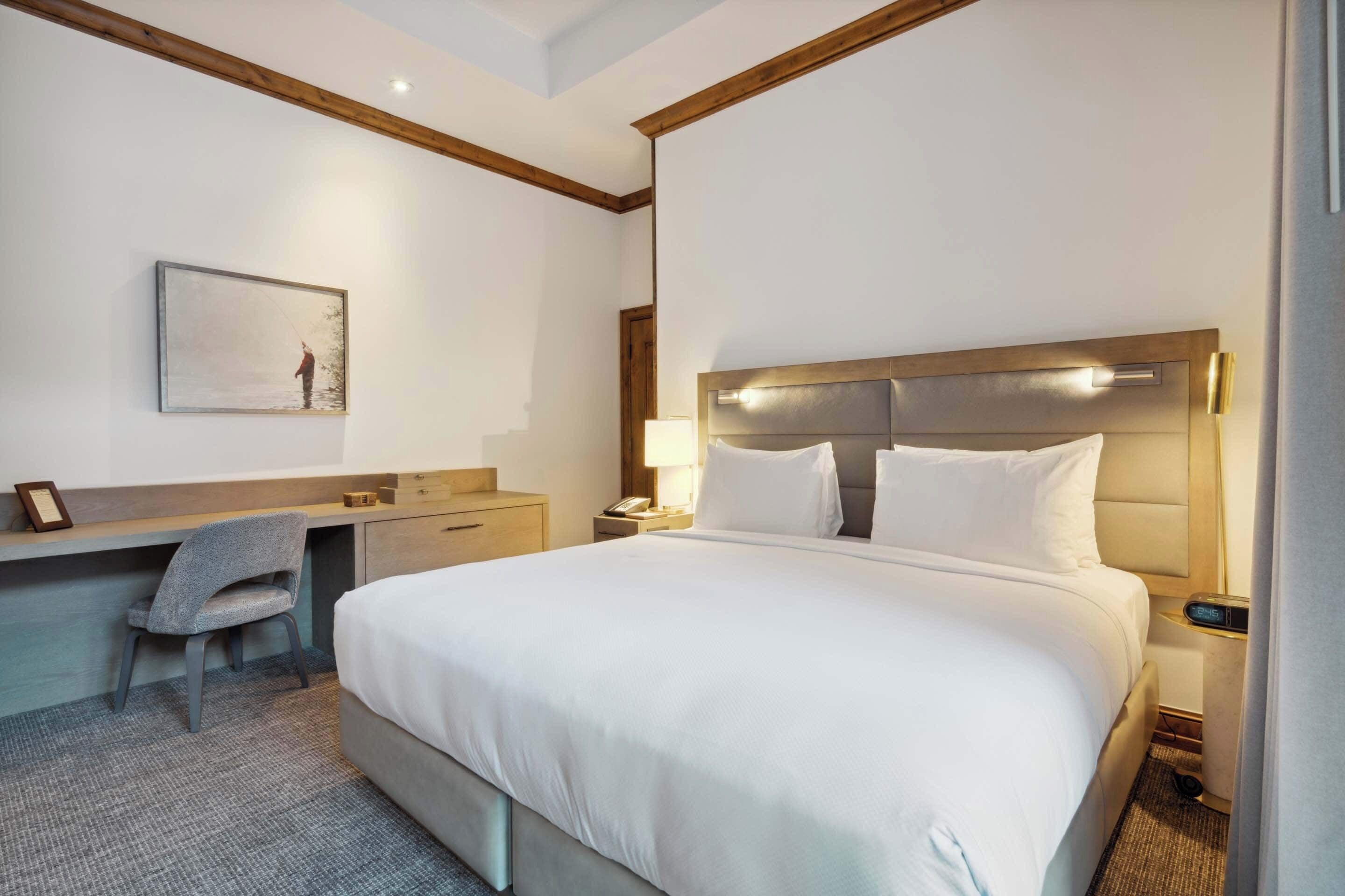A hotel room with a large bed, a desk with a chair, a bedside table, a lamp, and a framed picture on the wall.