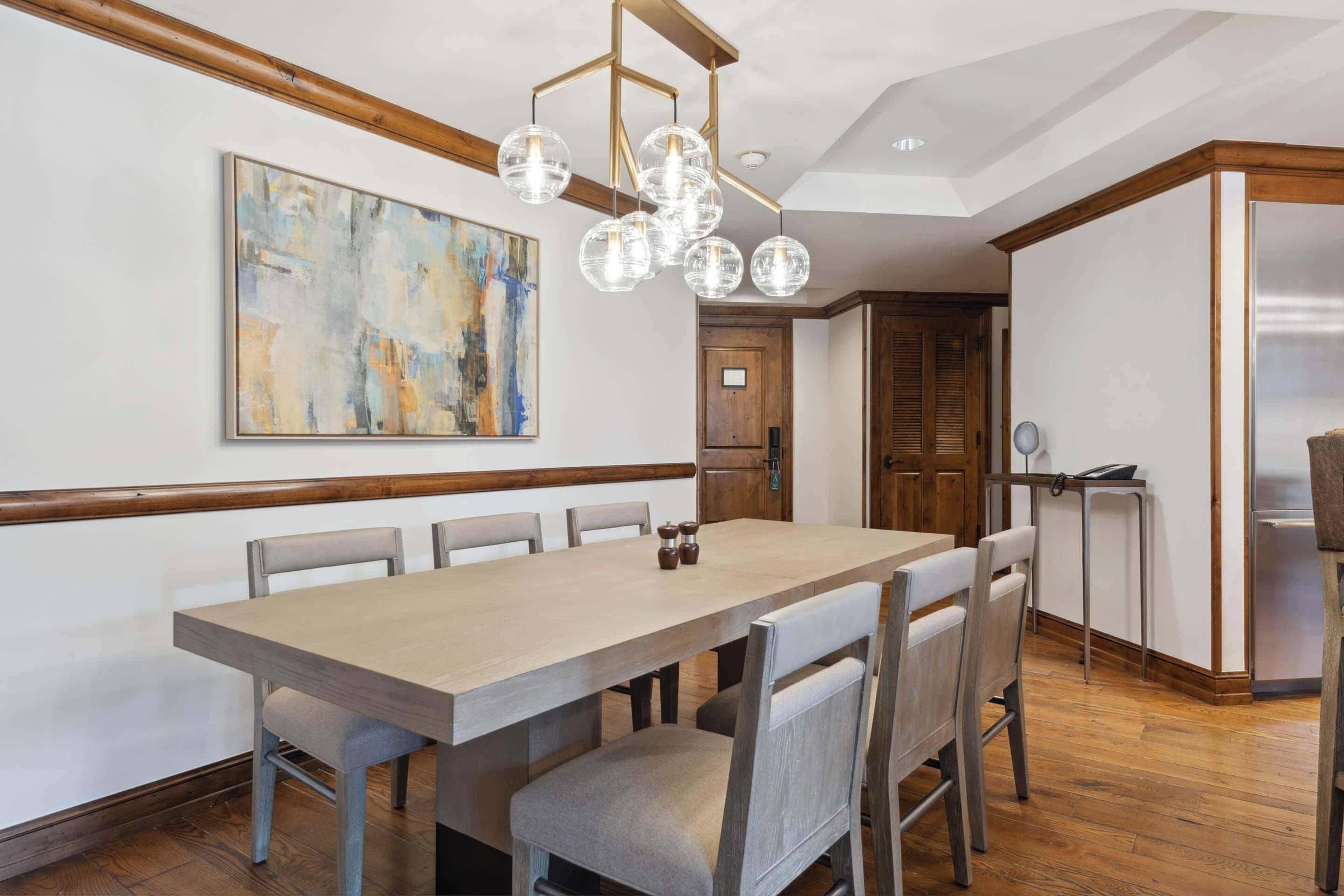 Un coin repas avec une longue table, six chaises, un lustre et un tableau accroché au mur.