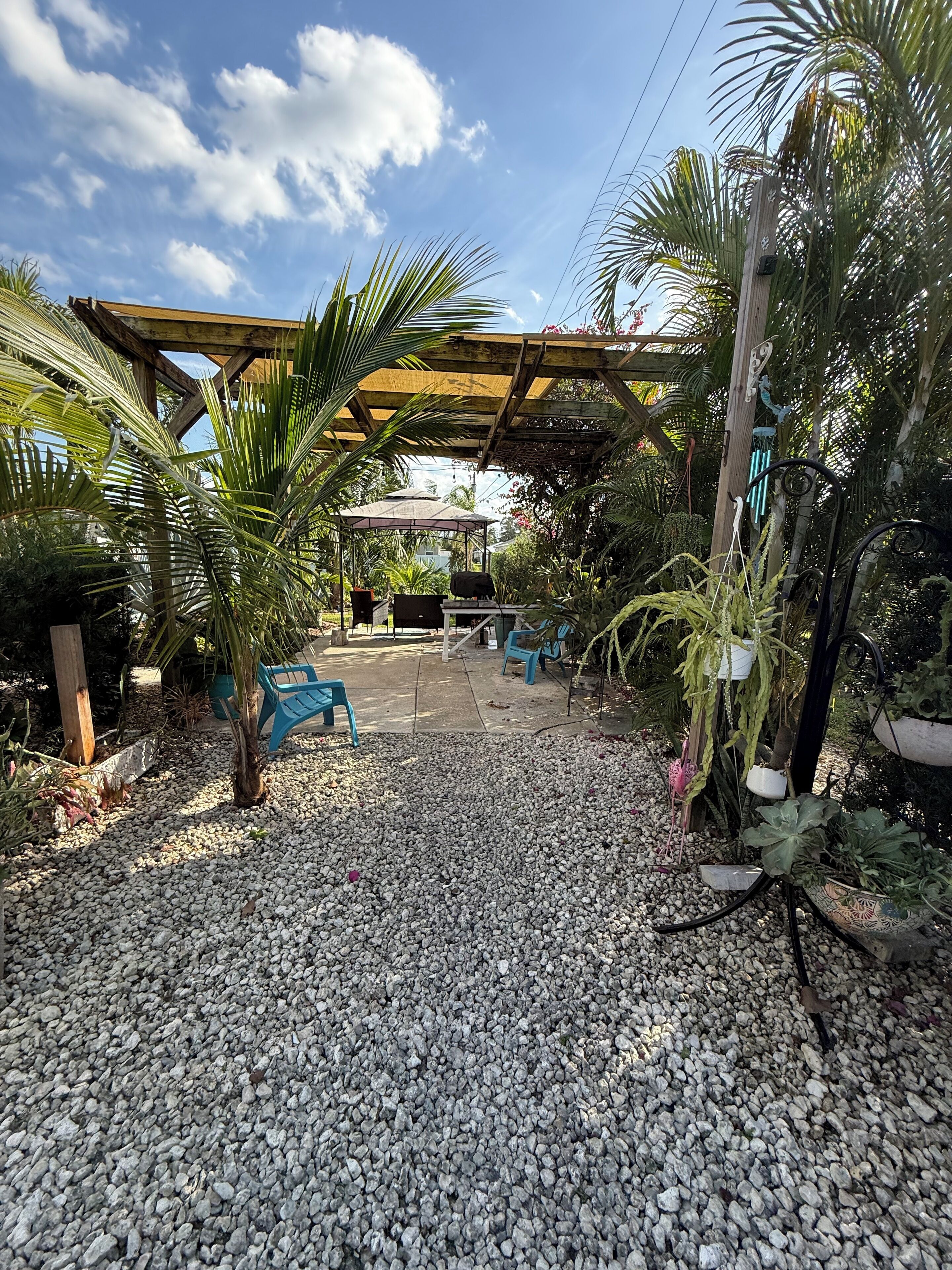 A covered outdoor seating area with a gravel floor, surrounded by various plants and trees.