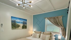 Une chambre à coucher avec un grand lit, un luminaire au plafond, un tableau représentant une scène de plage et une fenêtre avec des rideaux.