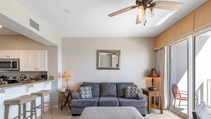 Ein Wohnzimmer mit grauem Sofa, zwei Barhockern, einem Couchtisch, einem Deckenventilator, einer Küche mit weißen Schränken und einer Glastür zum Balkon.