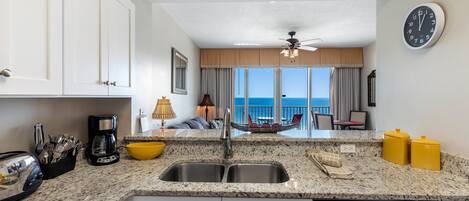 A kitchen with a granite countertop, white cabinets, a coffee maker, and a dishwasher. There is a view of the ocean through large windows.
