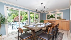 Un coin repas avec une table en bois dressée pour six personnes, entourée de chaises, d’un lustre et offrant une vue sur un jardin grâce à de grandes fenêtres.