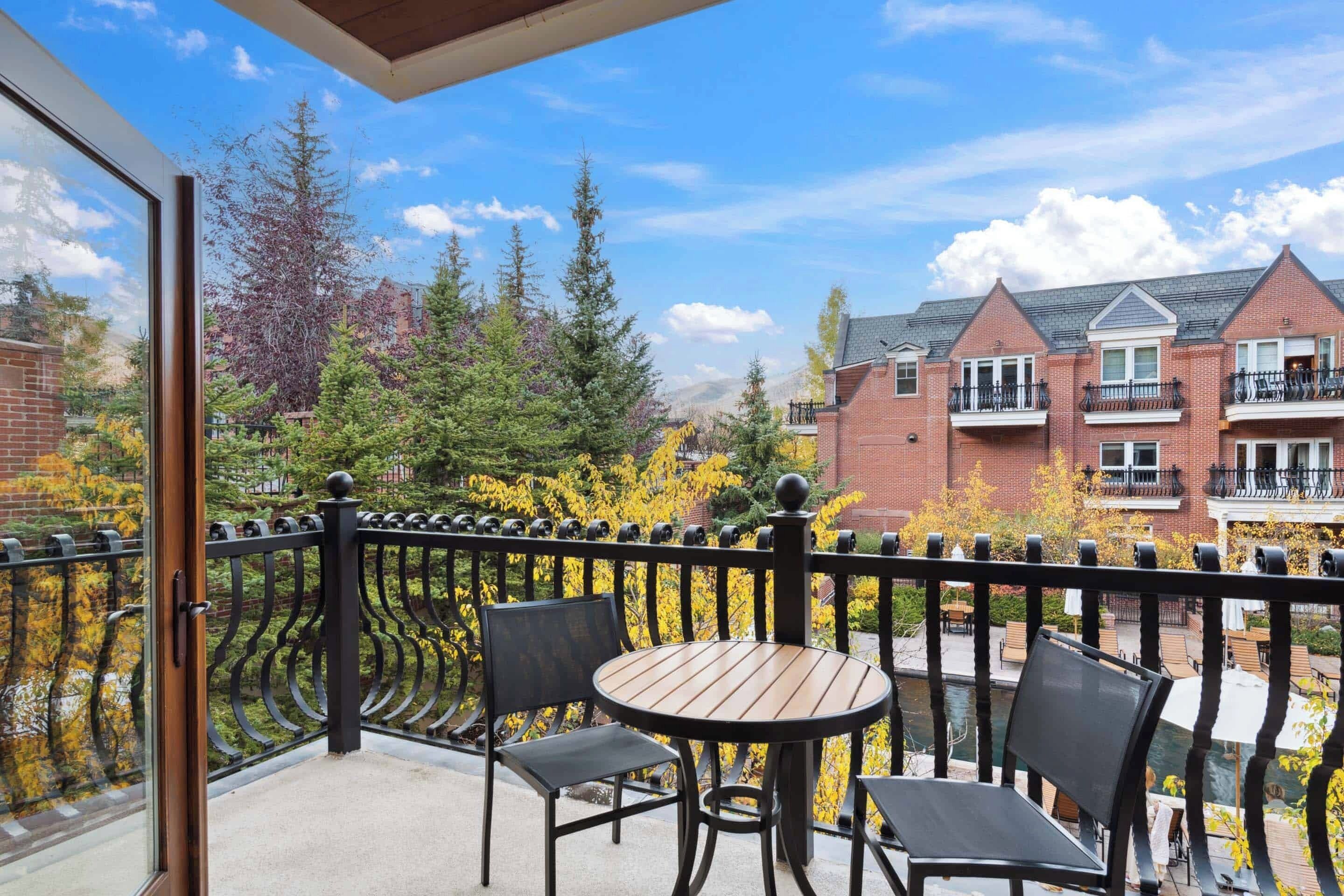 A balcony with a round table and two chairs, overlooking a courtyard with trees and a brick building in the background.