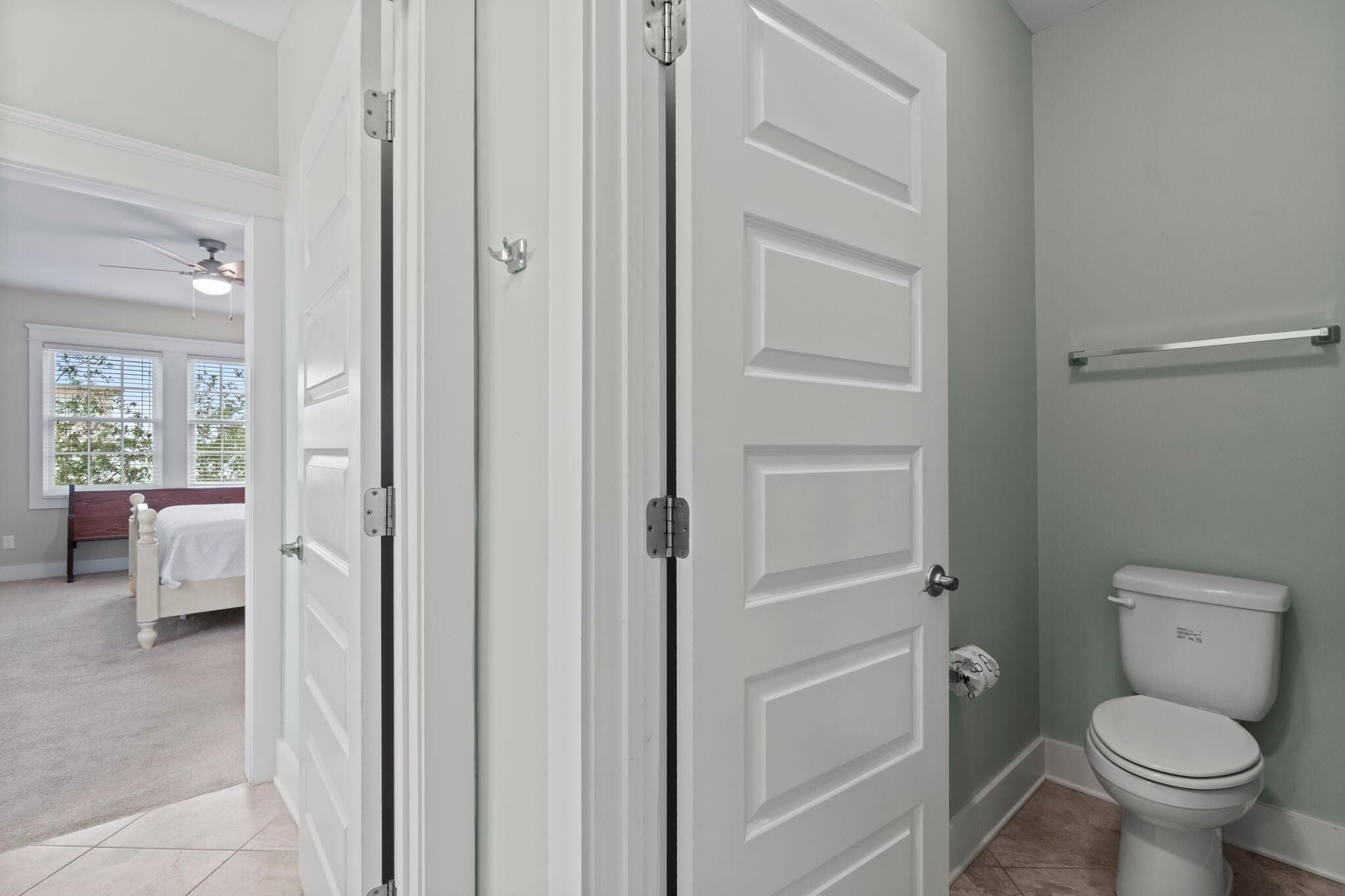 A bathroom with a toilet, a towel rack, and a door leading to a bedroom with a bed and a window.