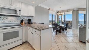 A modern kitchen with white cabinets, a granite countertop, and a built-in microwave. There is a dining area with a table and chairs, and a balcony with a view. - Phoenix Viii 1506 (Orange Beach)