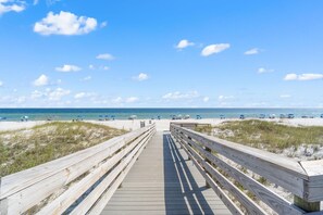 Ein hölzerner Steg führt zu einem Strand mit Dünen und blauem Himmel.