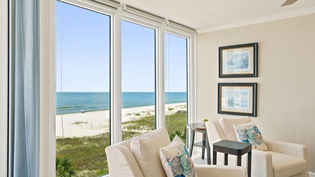A room with a large window overlooking a beach, a chair, a small table, and two framed pictures on the wall. - Seaspray West 503- Gulf Front (Pensacola)