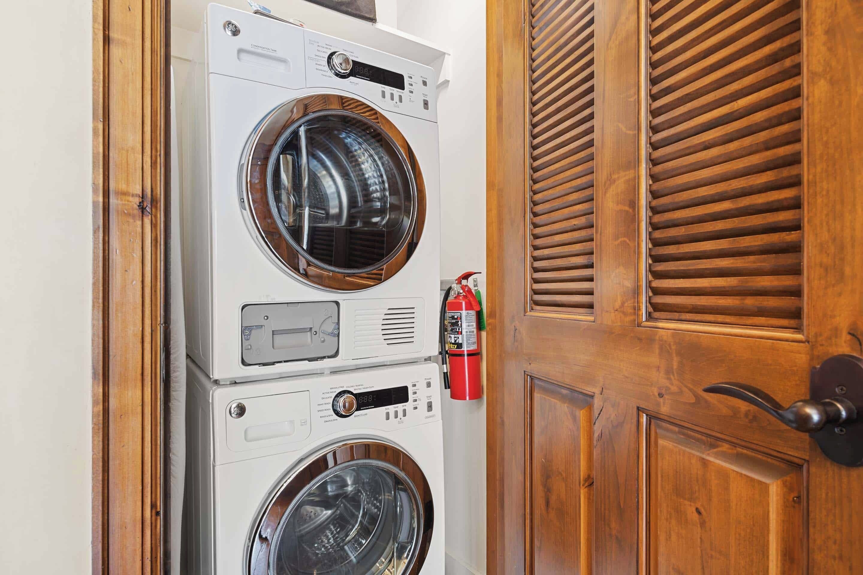 A stacked washer and dryer unit next to a wooden door with a fire extinguisher mounted on the wall.