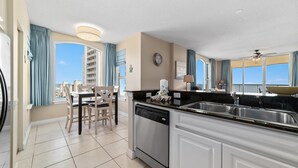 A modern kitchen with a dishwasher, sink, and dining area with chairs and a table. - Beach Colony East 7A (Pensacola)