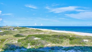 A coastal landscape with a beach, grassy dunes, and a small building with a flag. - Vista Del Mar 201 (Pensacola)