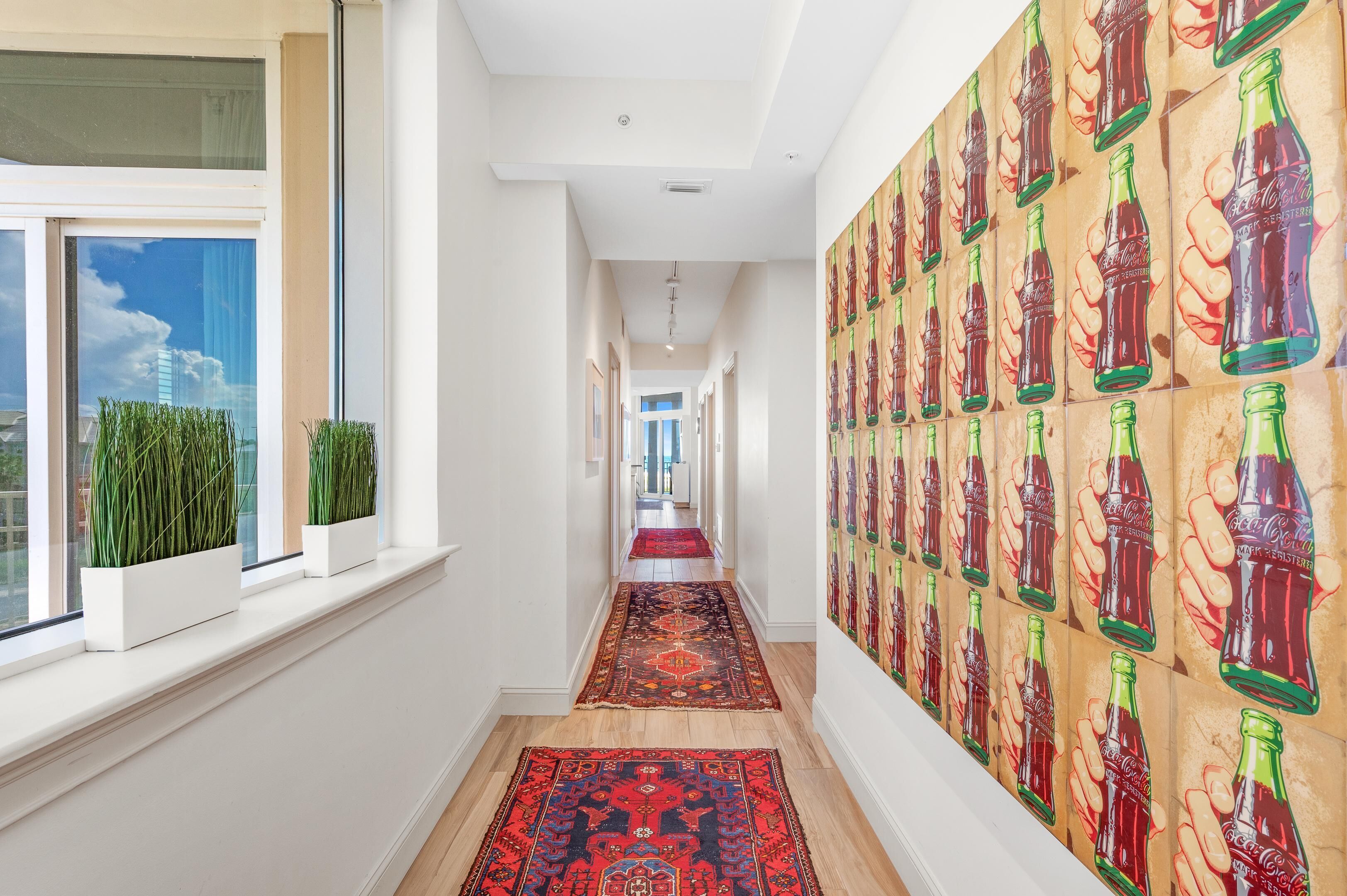 Un pasillo con alfombra roja, ventanas amplias con plantas y una pared decorada con murales de botellas Coca-Cola.