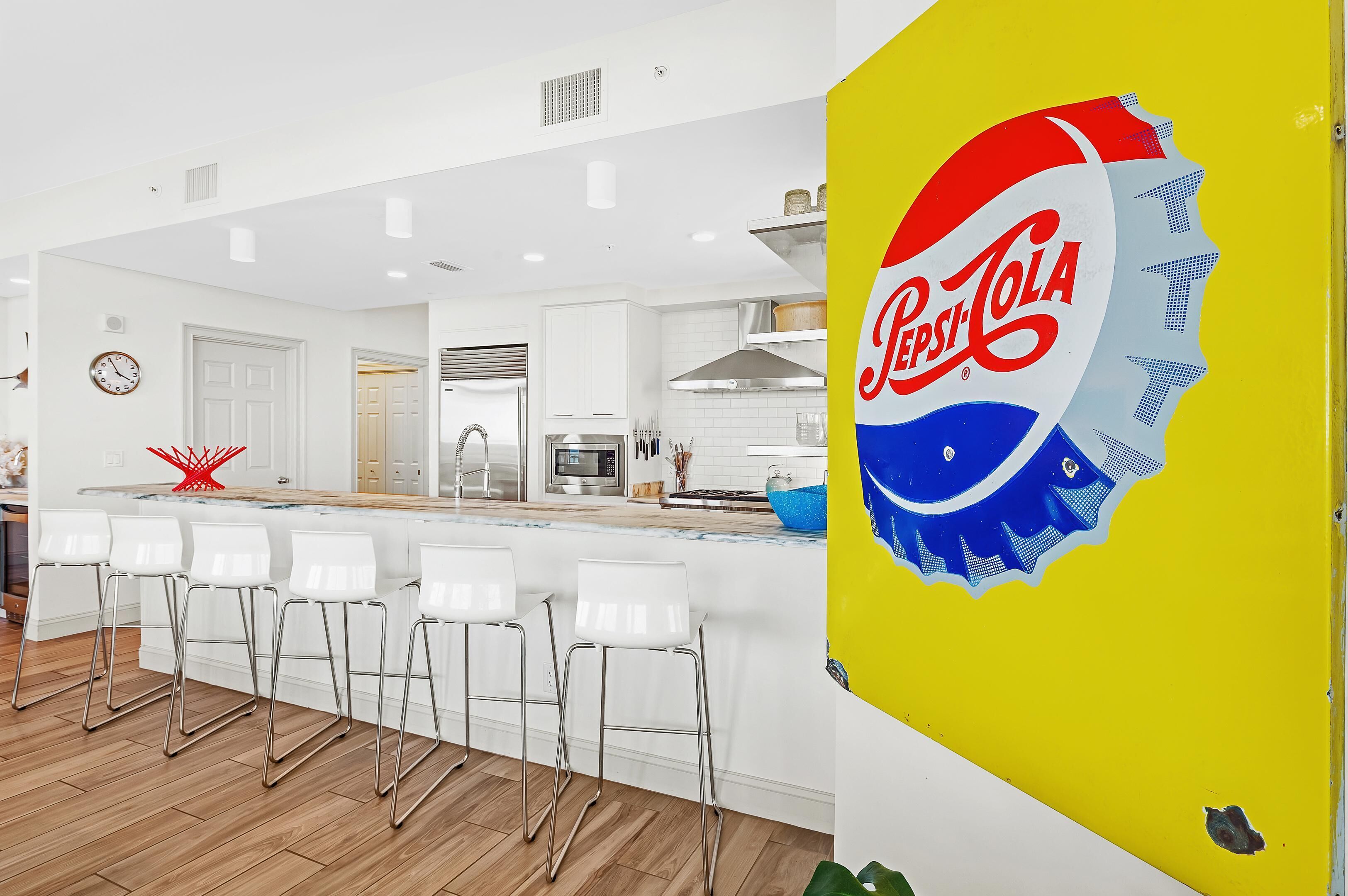 A modern kitchen with white cabinetry, a bar area with white stools, and a large Pepsi logo on a yellow door.