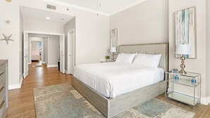 A bedroom with a bed, bedside tables, a nightstand, a ceiling fan, and a hallway leading to another room. - La Playa 301 (Pensacola)