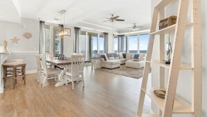 A spacious living room with a large window, a dining area with a wooden table and chairs, and a white bookshelf. - La Playa 301 (Pensacola)