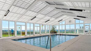 An indoor swimming pool with a clear view of the outdoors through large windows, a metal pool ladder, and a concrete floor. - Perdido Sun 712 (Pensacola)