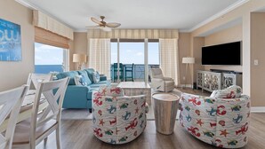 A living room with a blue sofa, two bean bag chairs with a sea-themed pattern, a ceiling fan, a TV, and a balcony with a view.