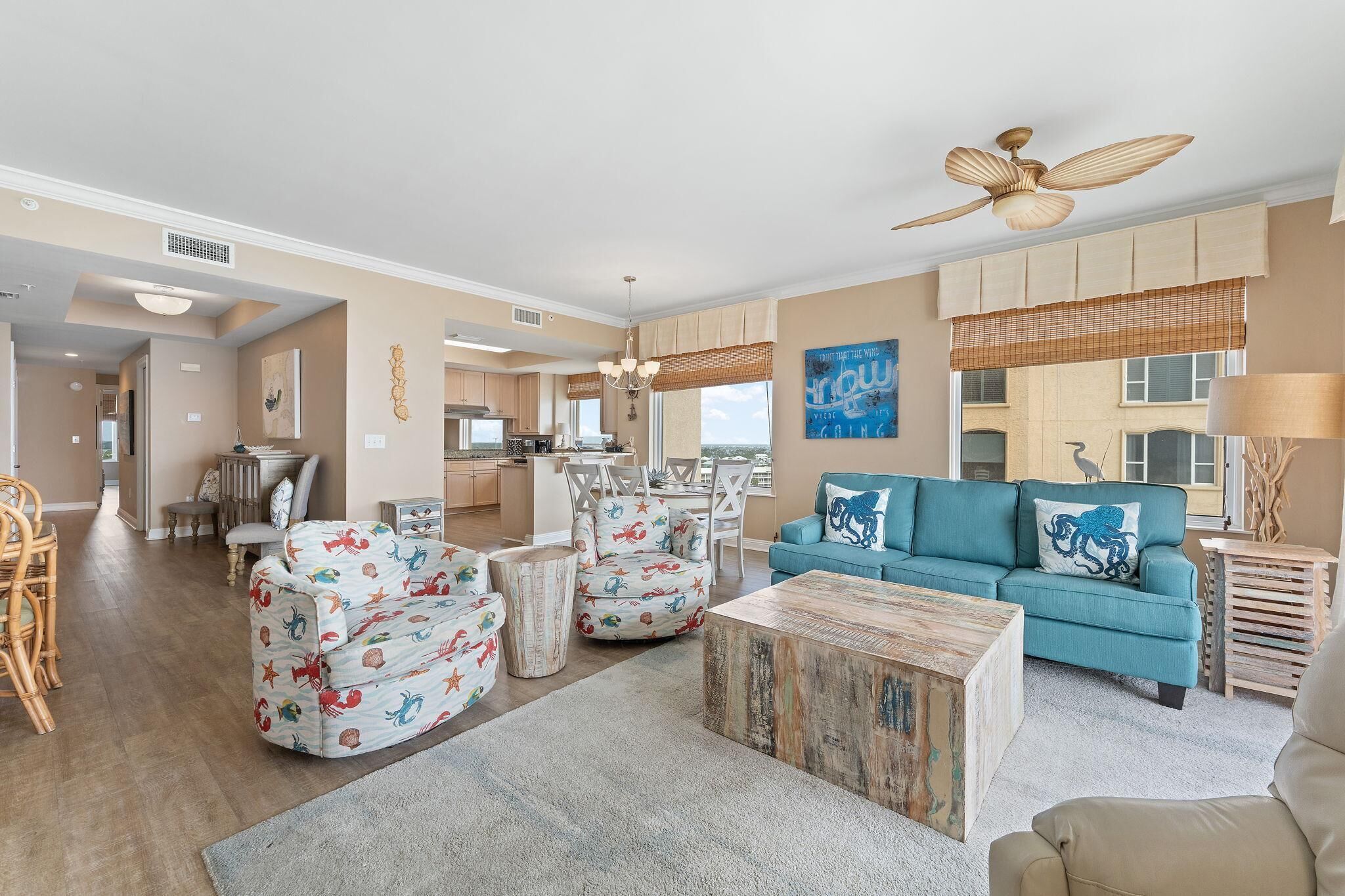 A spacious living room with a teal sofa, patterned armchairs, a wooden coffee table, and a dining area with a chandelier.