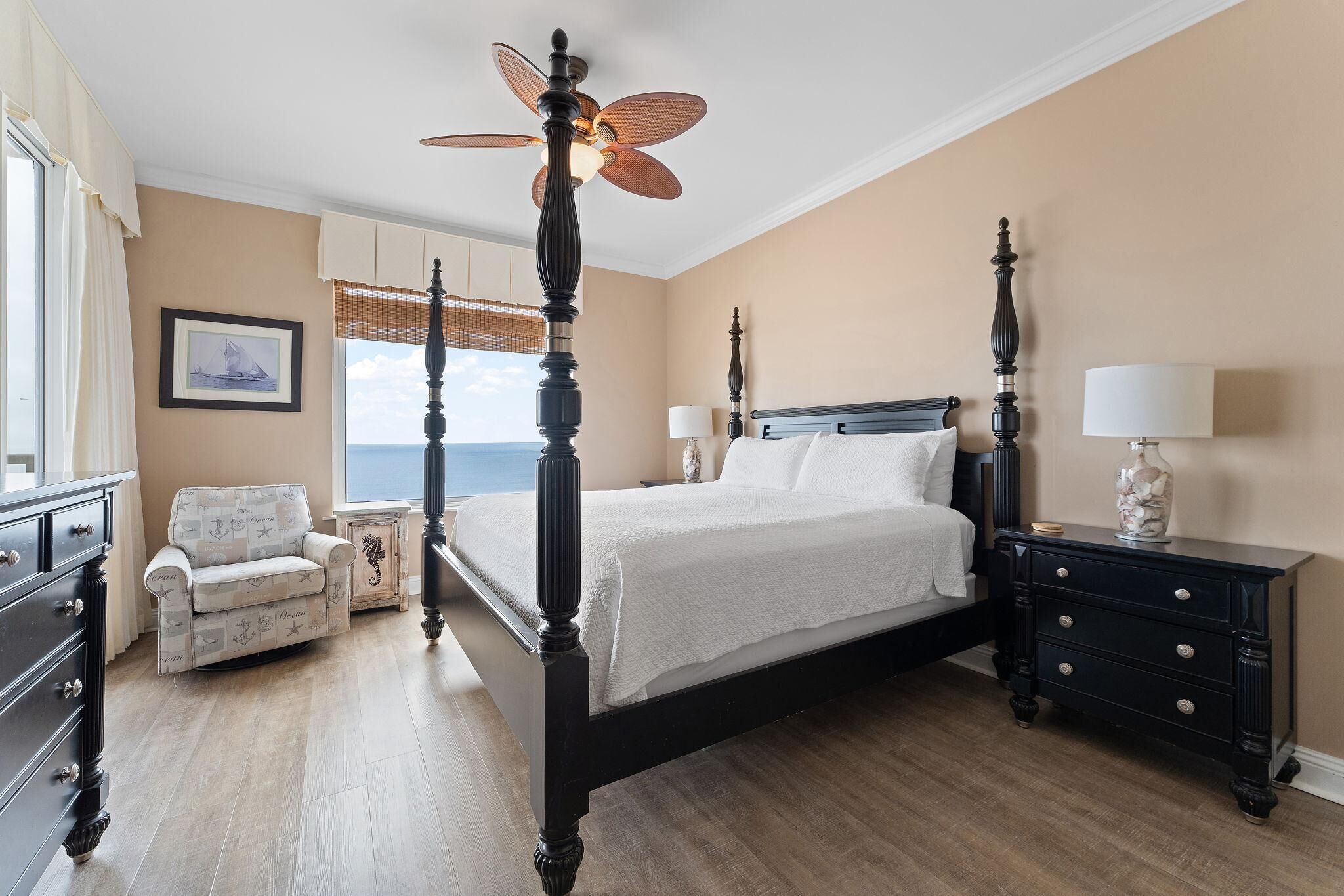 A bedroom with a four-poster bed, a chair, a nightstand, and a view of the sea.