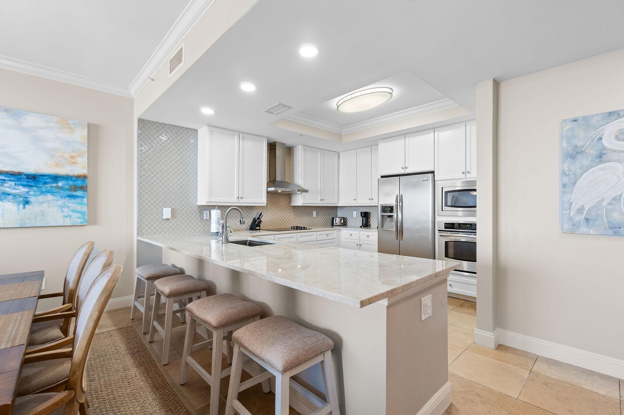 A modern kitchen with white cabinets, a large island with bar stools, stainless steel appliances, and artwork on the walls.
