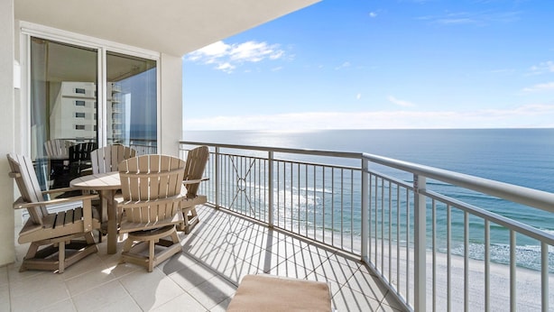 A balcony with a view of the ocean, a dining table with chairs, and a glass door leading to a room. - Indigo East 1602 (Pensacola)