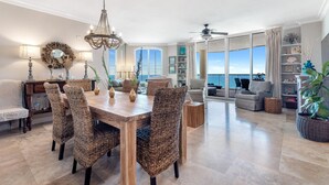 A spacious dining area with a wooden table and wicker chairs, a chandelier, and a view of the ocean through large windows. - Beach Colony East 16b-penthouse (Pensacola)