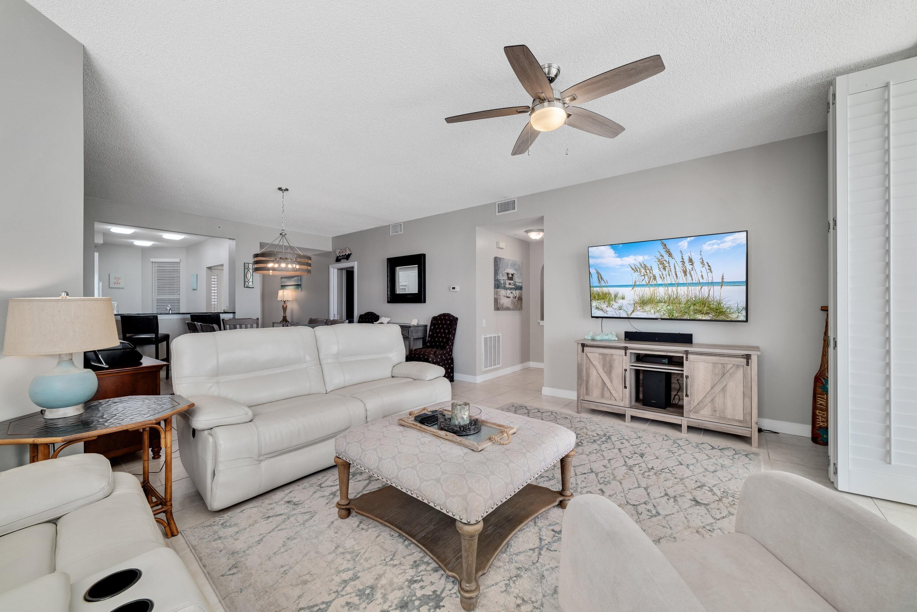 A spacious living room with a white leather sofa, a wooden coffee table, and a ceiling fan. A flat-screen TV is mounted on the wall, and there is a wooden cabinet with a TV stand.