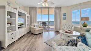 A spacious living room with a large TV, comfortable seating, a ceiling fan, and a view of the ocean through large windows.