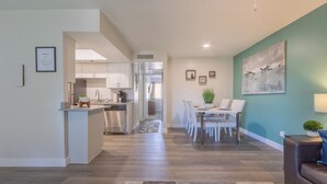 Una cocina moderna con armarios blancos, una zona de comedor con una mesa y sillas, y una sala de estar con un sofá marrón y un cuadro en la pared.