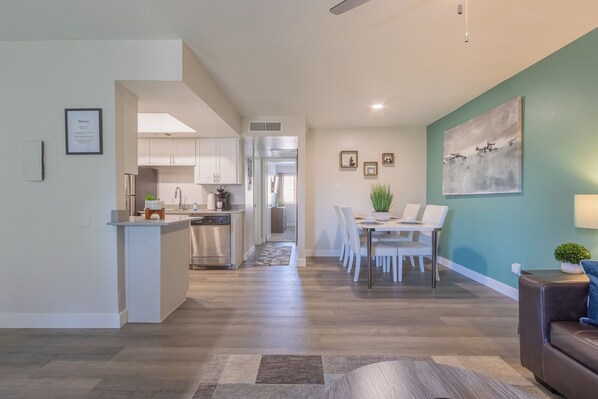 Una cocina moderna con armarios blancos, una zona de comedor con una mesa y sillas, y una sala de estar con un sofá marrón y un cuadro en la pared.