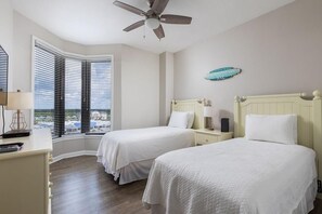 A hotel room with two beds, a ceiling fan, a window with blinds, a desk with a lamp, and a view of buildings outside. - Seachase 905c (Orange Beach)