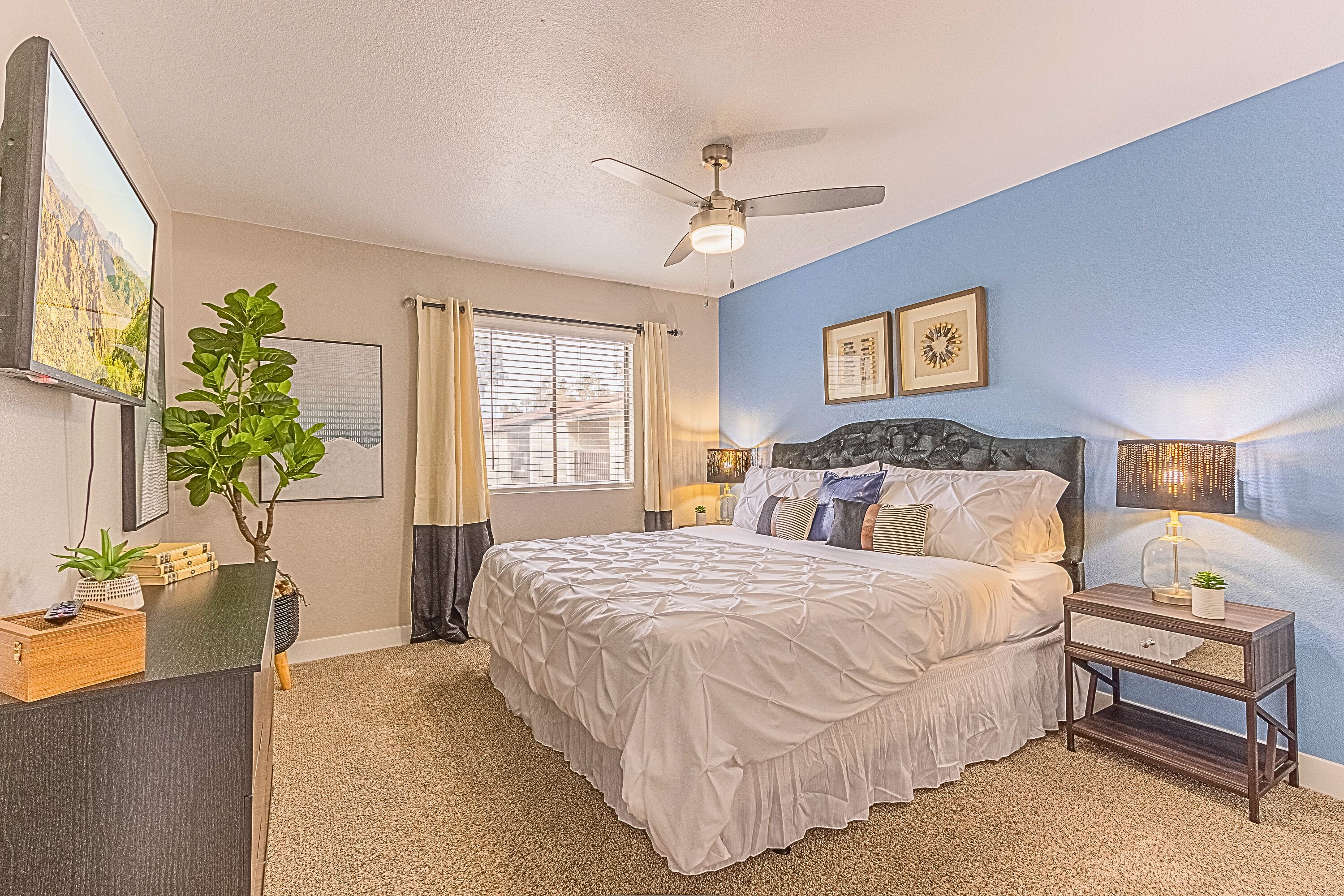 A bedroom with a bed, a ceiling fan, a window with curtains, a nightstand, a lamp, and a wall-mounted clock.
