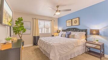 A bedroom with a bed, a ceiling fan, a window with curtains, a nightstand, a lamp, and a wall-mounted clock.