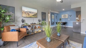 Een moderne woonkamer met een bank, salontafel en potplanten. Er is een eethoek met tafel en stoelen, en een keuken met een aanrecht en kasten.